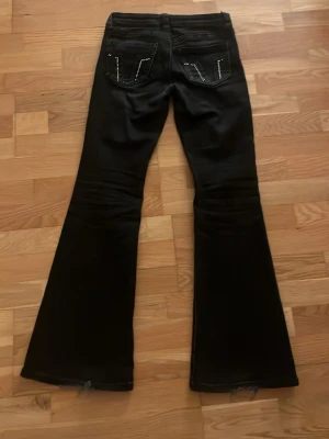 Svarta lowwaist bootcut jeans med nitar - Säljer ett par svarta bootcut jeans med snygga nitar på bakfickorna. Jeansen har en klassisk femficksmodell och utsvängda ben. Materialet är denim med stretch för extra komfort. Perfekta för dig som gillar en retro och trendig look. OBS!! Det saknas några nitar på fickorna (se bild) och är lite slitna längst ner på byxbenet (se bild)💗