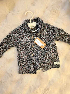 Blommig skjorta från Newbie - Söt skjorta från Newbie med blommigt mönster i vitt, rosa och grönt på svart botten. Skjortan har träknappar framtill och klassisk krage. Perfekt för dig som gillar detaljer och vill ha något unikt i garderoben. Nytt med tag storlek 62 för pojkar 
