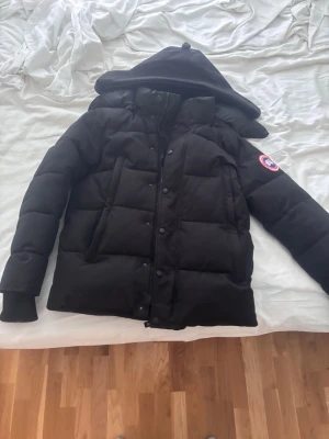 Svart pufferjacka från Canada Goose - Säljer en svart pufferjacka från Canada Goose med huva och klassisk logotyp på ärmen. Jackan har stora, rejäla paneler och stängs med dragkedja och knappar. Perfekt för kalla vinterdagar och riktigt varm. Passar dig som vill ha både stil och funktion.