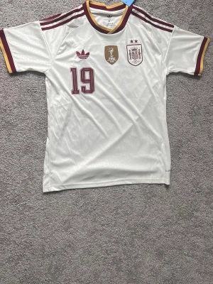  Spanien away fotbollströja lamine yamal  - Säljer en vit away Spanien fotbollströja från Adidas med vinröda detaljer, nummer 19 på bröstet och officiella FIFA-märket med lamine yamal bakom. Tröjan har korta ärmar, rund hals med gul och vinröd kant samt klassiska Adidas-ränder på axlarna. Perfekt för match eller träning.