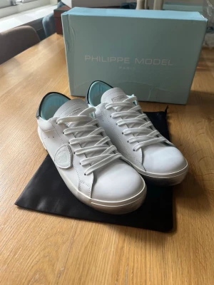 Vita Philippe Model sneakers - Stilrena vita sneakers från Philippe Model med svarta detaljer på hälen och ljusblå insida. Klassisk låg modell med vita skosnören och diskret logotyp på sidan. Tillverkade i slätt läder för en clean look.