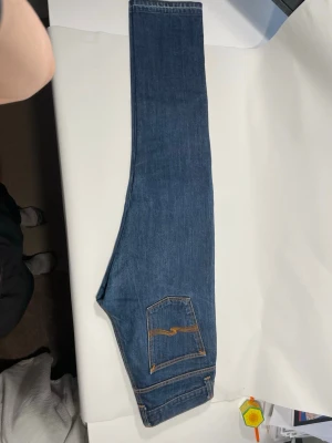 Blå jeans från Nudie Jeans - Säljer ett par klassiska blå jeans från Nudie Jeans med raka ben och femficksmodell. Jeansen har kontrastsömmar i orange och en snygg detalj på bakfickan. Tillverkade i slitstark denim, perfekta för en avslappnad stil. Nypris runt 2000 kr. Jeansen är selvage och riktigt fint skick. W32 L32