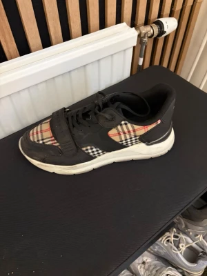 Burberry svart och rutig sneaker - Snygg sneaker från Burberry med klassiskt beige, rött och svart rutmönster. Skon har svarta detaljer i läder och textil, samt en vit sula. Tydlig Burberry-logga på remmen över tån. Perfekt för dig som vill sticka ut med en ikonisk design.