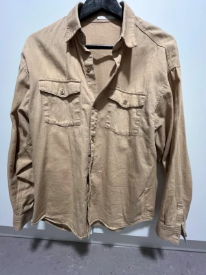 Beige overshirt i bomull - Stilren beige overshirt med två bröstfickor och knappar framtill. Skjortan har lång ärm och klassisk krage, tillverkad i mjuk bomull för en avslappnad look. Perfekt att bära öppen över en t-shirt eller stängd för en mer clean stil.