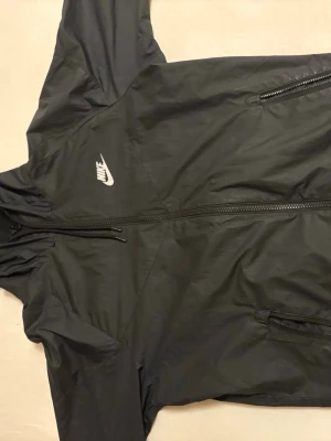 Svart vindjacka från Nike med huva - Svart vindjacka från Nike med vit logga på bröstet. Jackan har hel dragkedja, två fickor med dragkedja och en praktisk huva. Tillverkad i lätt och smidigt material som skyddar mot vind. Perfekt för en sportig och avslappnad stil.