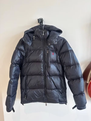 Mörkblå jacka från Polo Ralph Lauren - Snygg svart dunjacka från Polo Ralph Lauren med röd logga på bröstet. Jackan har huva, flera dragkedjeförsedda fickor och en glansig finish. Perfekt för kalla vinterdagar och riktigt stilren look . Skick 9/10, storlek S, nypris ≈ 6000kr