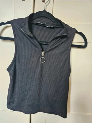 Topp lager 157 M L - Svart croppad ärmlös topp från 157 med ribbad struktur och hög krage. Toppen har en snygg dragkedja framtill med rund metallring som detalj. Perfekt att styla med jeans eller kjol för en cool look.  Kan även hämtas I Partille  E1