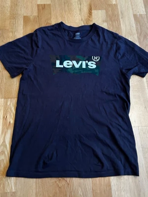 Svart Levi's t-shirt med camo-logga - Snygg svart t-shirt från Levi's med klassisk logga framtill i vitt mot en camouflagemönstrad bakgrund. T-shirten har rund hals och korta ärmar, perfekt till jeans eller shorts. Tillverkad i mjuk bomull för skön känsla hela dagen. Vid fundering hör av er 😁