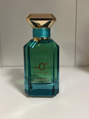 HERSH -0° Eau de Parfum 100 ml - Snygg parfymflaska från HERSH i turkost glas med guldfärgat lock och turkost band runt halsen. Flaskan har en modern, kantig design och rymmer 100 ml Eau de Parfum. Märkt med -0° Zero Degree Celsius för en fräsch känsla.