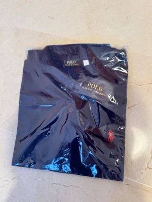 Marinblå skjorta från Polo Ralph Lauren - Snygg marinblå skjorta från Polo Ralph Lauren med klassisk krage och knappar framtill. Skjortan har den ikoniska röda logotypen broderad på bröstet och är tillverkad i ett mjukt material. Perfekt för en stilren look.