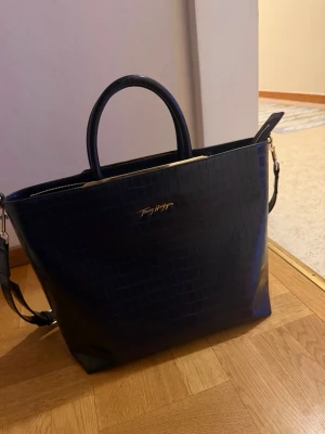 Tommy Hilfiger Svart handväska med krokoprägling - Stilren svart handväska med krokodilpräglat mönster och guldfärgade detaljer. Väskan har både handtag och en avtagbar axelrem för flexibel användning. Stängs med dragkedja och har en rymlig insida. Perfekt för dig som gillar klassisk men trendig design.