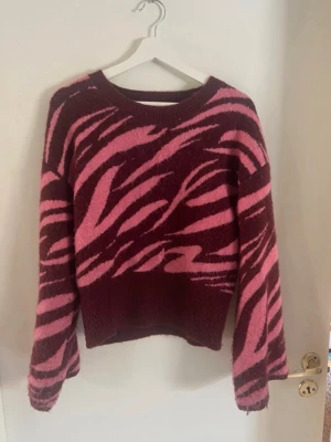 Vinröd och rosa stickad tröja Lindex - Stickad tröja från Lindex i vinrött med rosa zebramönster. Tröjan har rund halsringning, långa ärmar och ribbade muddar. Materialet är mjukt och mysigt, perfekt för kyligare dagar. Snygg och trendig design som sticker ut.