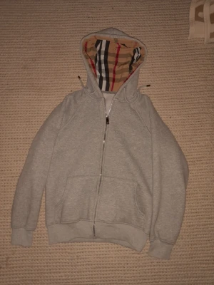 Grå burberry hoodie  - Snygg grå hoodie med dragkedja och känguruficka. Huvan har ett klassiskt rutigt mönster i beige, svart, vitt och rött som ger en unik touch. Perfekt för dig som vill ha en stilren men ändå utstickande look. Mjuk bomullskänsla och ribbade muddar.