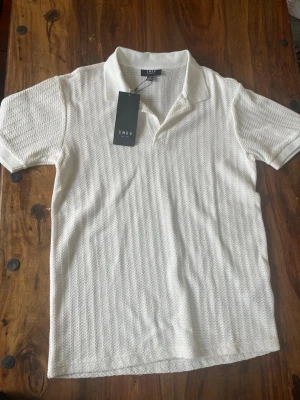 Vit piké t-shirt från SMOG - Säljer en fräsch vit piké t-shirt från SMOG med korta ärmar och klassisk krage. Tröjan har ett diskret strukturerat mönster i tyget och är tillverkad i mjuk bomull. Perfekt för en clean och stilren look.