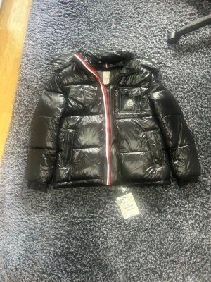 Svart dunjacka från Moncler - Snygg svart dunjacka från Moncler med glansig finish och klassisk logotyp på bröstfickan. Jackan har huva, dragkedja med rödvit detalj och flera praktiska fickor. Perfekt för kalla vinterdagar och riktigt trendig look.