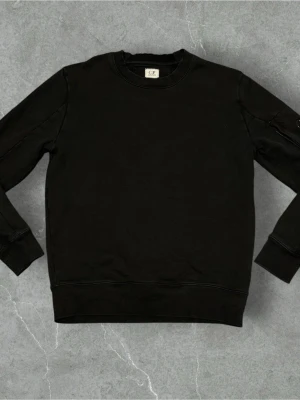 Svart sweatshirt från C.P. Company - En stilren svart sweatshirt från C.P. Company med rund halsringning och ribbade muddar vid ärmslut och nederkant. Tröjan har en diskret ficka med dragkedja på ena ärmen som ger en cool detalj. Perfekt för en avslappnad look.
