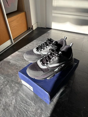 Svarta chunky sneakers med meshdetaljer - Säljer ett par svarta chunky sneakers med coola meshdetaljer och grå inslag. Skorna har en robust sula, svarta och vita skosnören samt dragflikar bak och fram. Perfekta för dig som gillar streetwear och vill ha en modern look.
