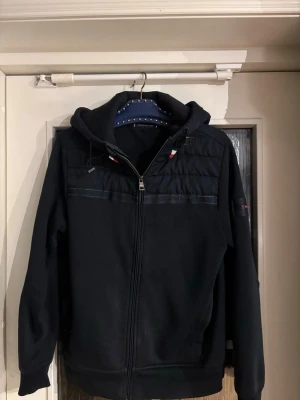 Tommy Hilfiger Cardigan - Marinblå cardigan jacka med huva och dragkedja framtill. Jackan har quiltade detaljer upptill, justerbar snörning vid huvan och ribbade muddar. Perfekt för dig som gillar en stilren och funktionell look.