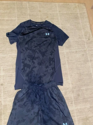 Under Armour träningsset camo svart - Snyggt träningsset från Under Armour med t-shirt och shorts i svart med diskret camomönster. Båda plaggen har Under Armour-logga i neon. Materialet är lätt och andas, perfekt för träning eller gym. T-shirten har kort ärm och shortsen har resår i midjan. Settet är andvänd runt 3 gånger så alltså ny skick, inget fel med de inga deffekter. Priset är spikat. Byxornas size är XS och tshirten S