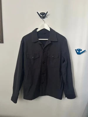Svart overshirt med bröstfickor ( uniqlo ) - Stilren svart overshirt med knäppning framtill och två bröstfickor med knapp. Klassisk krage och långa ärmar. Perfekt lager-på-lager-plagg för en avslappnad look. Materialet ser ut att vara bomull eller en bomullsblandning.
