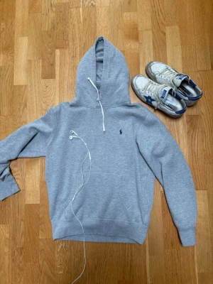 Grå hoodie från Polo Ralph Lauren - Snygg grå hoodie från Polo Ralph Lauren med klassisk huva och dragsko. Tröjan har det ikoniska lilla logobroderiet på bröstet och är gjord i mjukt bomullsmaterial. Perfekt för en chill och stilren look. 