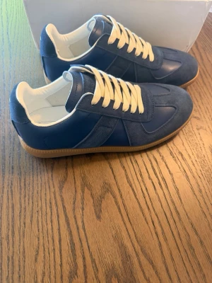 Mason Margiela Gats - Snygga blå sneakers med både mocka och släta paneler, vita snören och vit insida. Skorna har en klassisk gummisula i brunt och låg profil. Perfekta för dig som gillar stilrena och tidlösa sneakers med en modern touch.