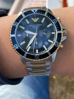 Emporio Armani World Explorer klocka silver/guld - Emporio Armani World Explorer klocka med metallarmband i silver och guldfärgade detaljer. Urtavlan är svart med kronograf och tydliga indexmarkeringar. Klassisk rund form och robust design, perfekt för dig som gillar stilrena accessoarer. Klockan är i nyskick och skickas med original lådan!