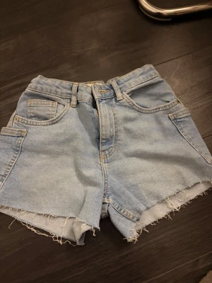 Ljusblå denimshorts med rå kant - Säljer ett par ljusblå denimshorts med hög midja och rå, fransig kant nedtill. Shortsen har klassiska fem fickor, bälteshällor och dragkedja framtill. Perfekta för sommaren och ger en avslappnad vibe.