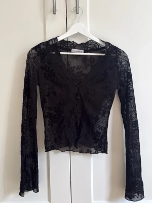 Svart spetsblus med v-ringning - Säljer en svart, transparent spetsblus med v-ringning och långa ärmar. Blusen har ett blommigt mönster och knäppning framtill. Perfekt för dig som vill ha en edgy och trendig look💗
