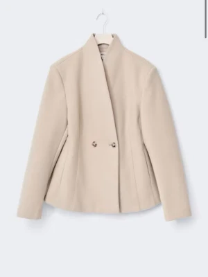 Beige kavaj med hög krage - Stilren beige kavaj med hög krage och dubbelknäppning framtill. Kavajen har en figurnära passform och långa ärmar. Tillverkad i ett slätt tyg som ger en elegant look. Perfekt för dig som gillar minimalistisk och modern stil. Slutsåld på hemsidan i alla storlekar 