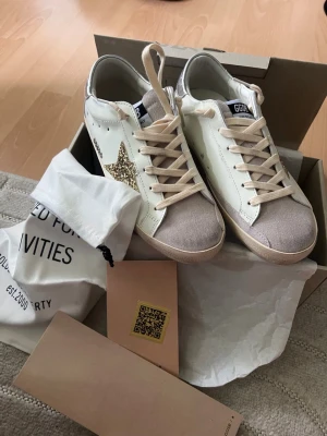 Vita sneakers med glitterstjärna - Snygga vita sneakers med beige detaljer och en glittrig guldstjärna på sidan. Skorna har beige snören, grå mockaliknande tå och vit sula. Märket GGDB syns på plösen. Perfekta för dig som vill sticka ut med en unik detalj.