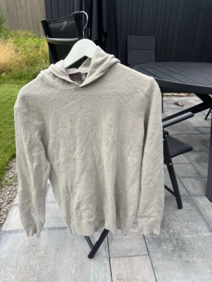 Beige hoodie från Oscar Jacobson - Säljer en stilren beige  hoodie från Oscar Jacobson i mjukt material. Hoodien har en klassisk huva och ribbade muddar vid ärmslut och nederkant. Perfekt för dig som gillar enkel och clean stil. Tillverkad i Italien.