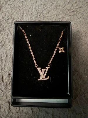 Guldhalsband med LV-hänge - Snyggt halsband i guldfärgad metall med ett stort LV-hänge täckt av små glittrande stenar. Kedjan har även en liten blomformad detalj på sidan. Perfekt accessoar för dig som gillar statement-smycken och vill sticka ut.