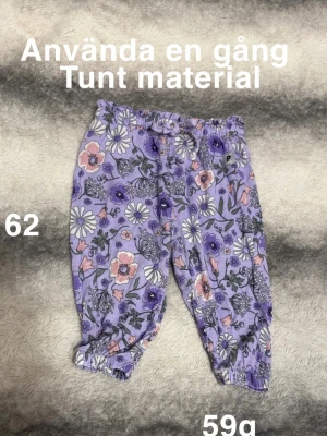Lila blommiga byxor med resår - Tunna byxor i lila med blommigt mönster i vitt, rosa och grönt. Byxorna har resår i midjan och vid bensluten för en skön och flexibel passform. Perfekta för dig som gillar färg och mönster.🌸 Använda en gång. Tunt material. Storlek 62