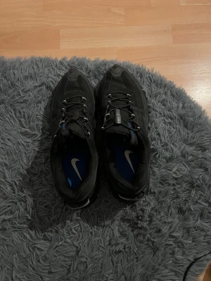Svarta Nike sneakers med blå detalj - Säljer ett par svarta Nike sneakers med blå innersula och vita detaljer på snörningen. Skorna har en chunky sula och lågt skaft, samt reflekterande detaljer på hälen. Perfekta för dig som gillar sportig och clean stil.