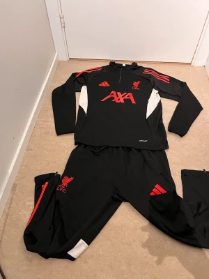 Liverpool svart träningsset Adidas - Säljer ett svart träningsset från Adidas med Liverpool FC-logga. Setet består av en långärmad tröja med halv dragkedja och matchande byxor. Röda detaljer och loggor samt vita paneler på sidorna. Perfekt för träning eller uppvärmning.