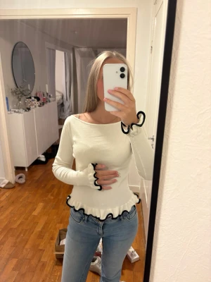 Vit långärmad topp med volang - Söt vit långärmad topp med volangkant och svarta vågiga detaljer vid ärmslut och midja. Toppen har en rund halsringning och är figurnära i modellen. Perfekt för dig som gillar en feminin och trendig stil.