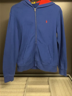 Blå Ralph lauren hoodie - Snygg blå hoodie med dragkedja framtill och röd insida i huvan. Klassisk design med två fickor på magen och diskret logga broderad på bröstet. Tillverkad i mjukt bomullsmaterial som känns skönt mot huden. Perfekt för en avslappnad och stilren look.