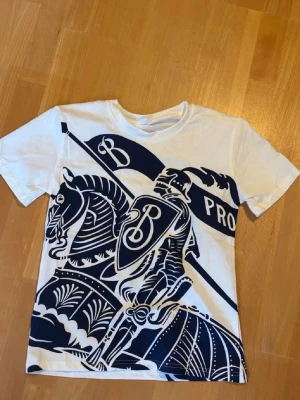Burrbery T shirt - Säljer en vit t-shirt med en stor, mörkblå grafisk print av en riddare på häst och flagga på framsidan. T-shirten har rund hals och korta ärmar. Materialet känns som bomull och passformen är normal.