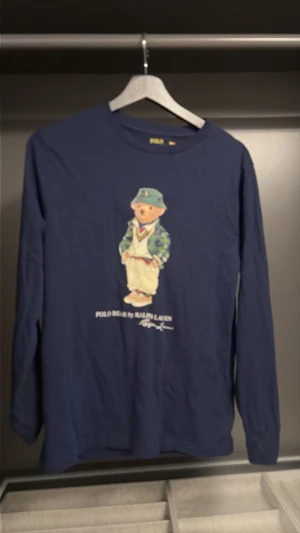 Blå långärmad t-shirt Polo Bear - Mörkblå långärmad t-shirt från Polo Ralph Lauren med ikoniskt Polo Bear-tryck på bröstet. Rund halsringning och mjukt bomullsmaterial. Perfekt för en avslappnad och trendig stil.