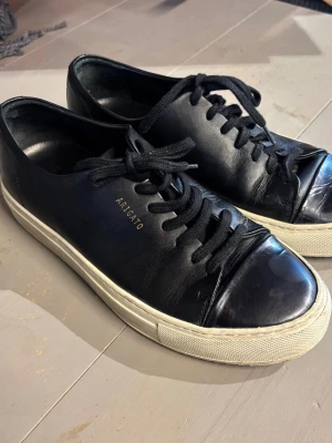Svarta sneakers från Axel Arigato - Säljer ett par snygga Arigato-sneakers i svart skinn med glansig tå. Skorna är i fint använt skick med lite slitningar framtill (se bilder).  Köpta på Sellpy men var tyvärr för stora för mig. De såldes som storlek 36, men jag skulle säga att de passar mer som en ca 38. Det finns ingen storleksmärkning som jag kan hitta.☺️