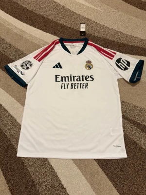 Real Madrid 26-27 hemmatröja  - Real Madrid 26-27 hemmatröja   Storlek: XXL Namn och nummer: MBAPPÉ #10 Badge: Champions League  Beskrivning: Tröjan har fel font på baksidan och fel UEFA Foundation badge på sidan under bollen.  Säljs av ICEKIT icekit.se