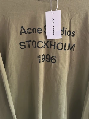 Beige långärmad topp Acne Studios - Cool beige långärmad topp från Acne Studios med tryck 'STOCKHOLM 1996' framtill. Oversized fit och mjukt bomullsmaterial gör den perfekt för en avslappnad look. Snyggt tryck i svart ger en trendig vibe.
