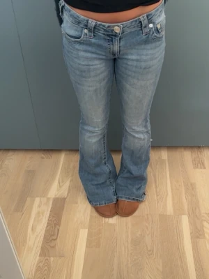 True religion jeans med Rosa detail - Säljer ett par ljusblå bootcut jeans med snygga slitningar och klassiska fem fickor. Jeansen har en låg midja och dekorativa sömmar på bakfickorna. Perfekta för en avslappnad och trendig look. Knappt använda då dom är förstora💕