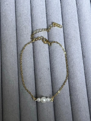 Guldfärgat armband med pärlor från  - Snyggt armband i guldfärgad metall med tre vita pärlor i mitten. Kedjan är 18k guldplaterat och har ett justerbart lås med en liten bricka där märket står ingraverat. Perfekt för dig som gillar stilrena och eleganta accessoarer.