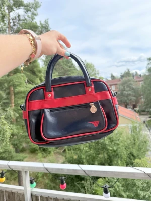 Svart och röd retro handväska y2k vines - Cool svart handväska med röda detaljer och rundad shape. Väskan har dubbla handtag, en framficka med dragkedja och dekorativt emblem. Tillverkad i glansigt konstläder som ger en vintagekänsla. Perfekt för dig som gillar färgstarka accessoarer. Aldrig använd! Supersnygg. Har även en innerficka!