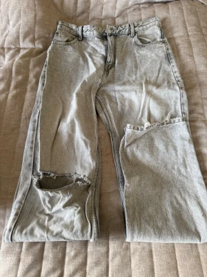Gråa mid waist jeans från Bershka - Säljer ett par ljusgrå mid waist jeans från Bershka med rak passform. Jeansen har slitningar och hål på ena benet för en trendig, avslappnad look. Klassisk femficksmodell och bälteshällor. 
