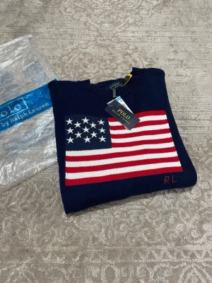 Marinblå stickad tröja Polo Ralph Lauren - Snygg marinblå stickad tröja från Polo Ralph Lauren med amerikansk flagga i rött, vitt och blått på bröstet. Tröjan har rund halsringning, ribbade muddar och RL-broderi. Perfekt för dig som gillar klassisk och preppy stil. Den passar S men även M
