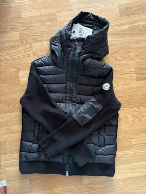 Svart cardigan med huva från Moncler - Snygg svart hybridjacka från Moncler med vadderad kropp och stickade ärmar. Jackan har huva med dragsko, två sidofickor med dragkedja och Moncler-logga på ärmen. Perfekt för lager-på-lager och streetwear-stil. Pris kan diskuteras 