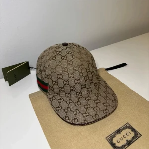 Gucci beige keps med GG-mönster - Snygg beige keps från Gucci med klassiskt GG-monogram över hela kepsen. Kepsen har en böjd skärm och en grön-röd rand på sidan. Tillverkad i canvasmaterial och levereras med originalpåse från Gucci. Perfekt för dig som gillar exklusiva accessoarer.